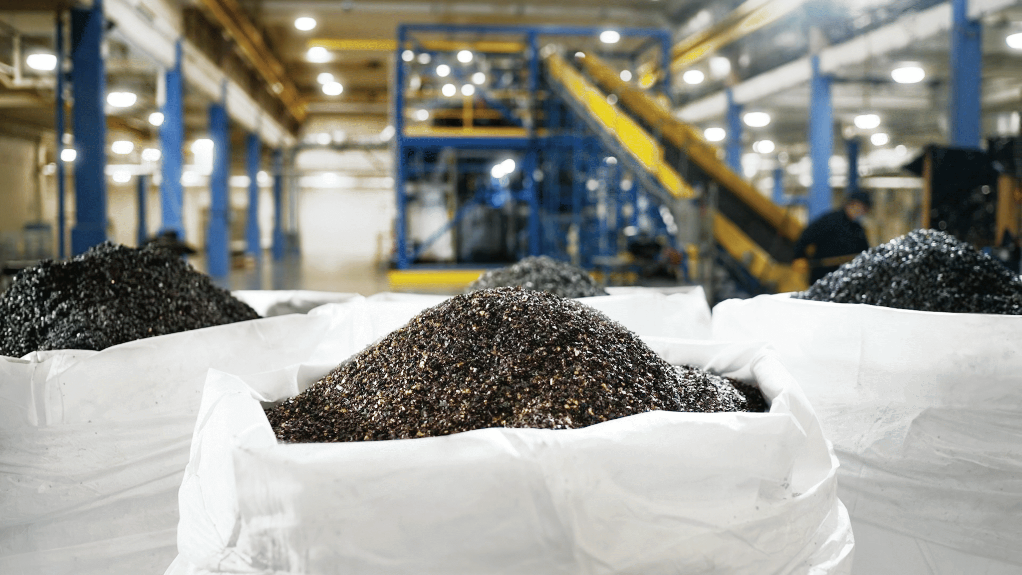 Three bags of black powder (black mass) at the forefront of the image. Conveyor belt is in the background of the image.