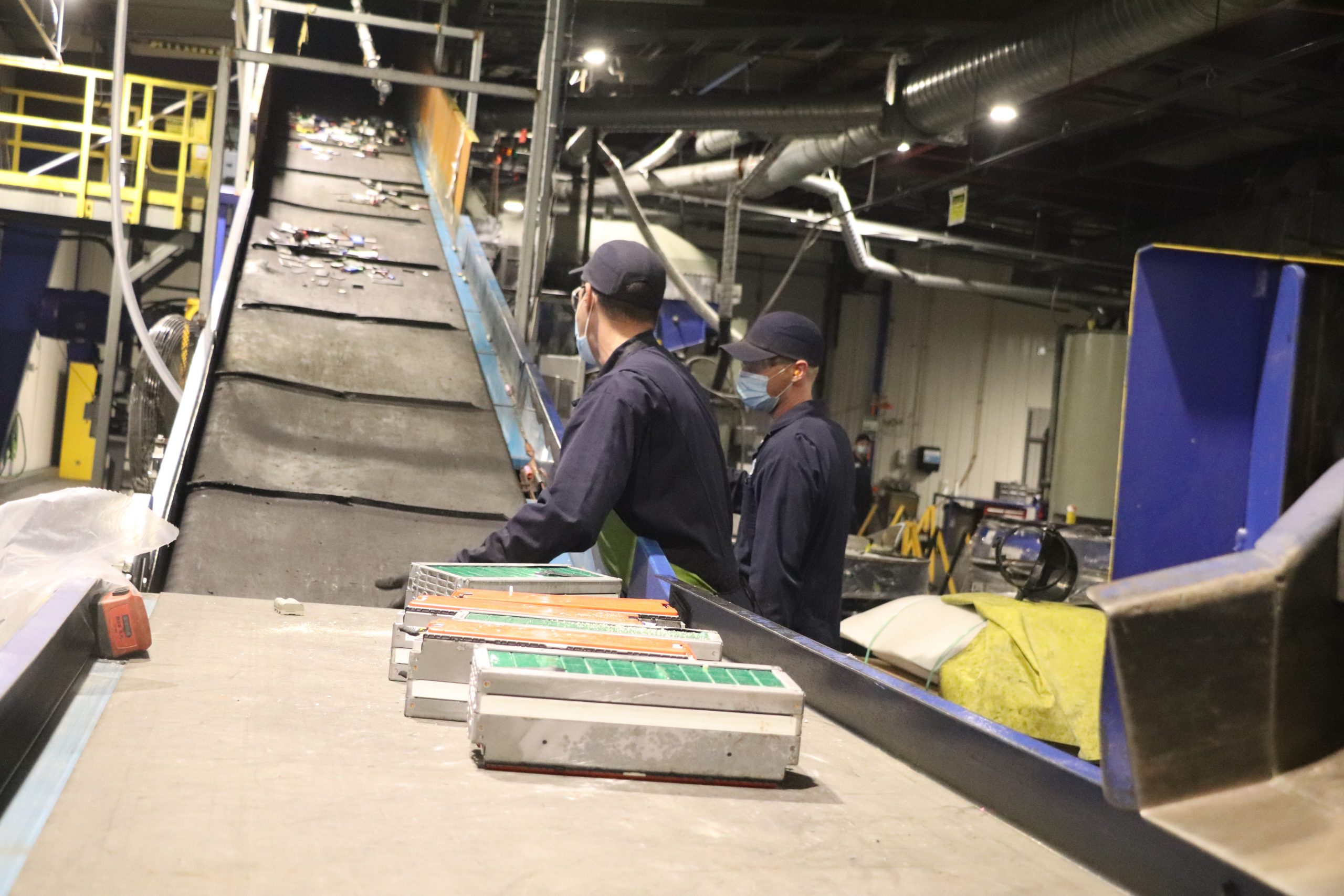 Two Li-Cycle employees wearing safety gear loading a conveyor belt with batteries